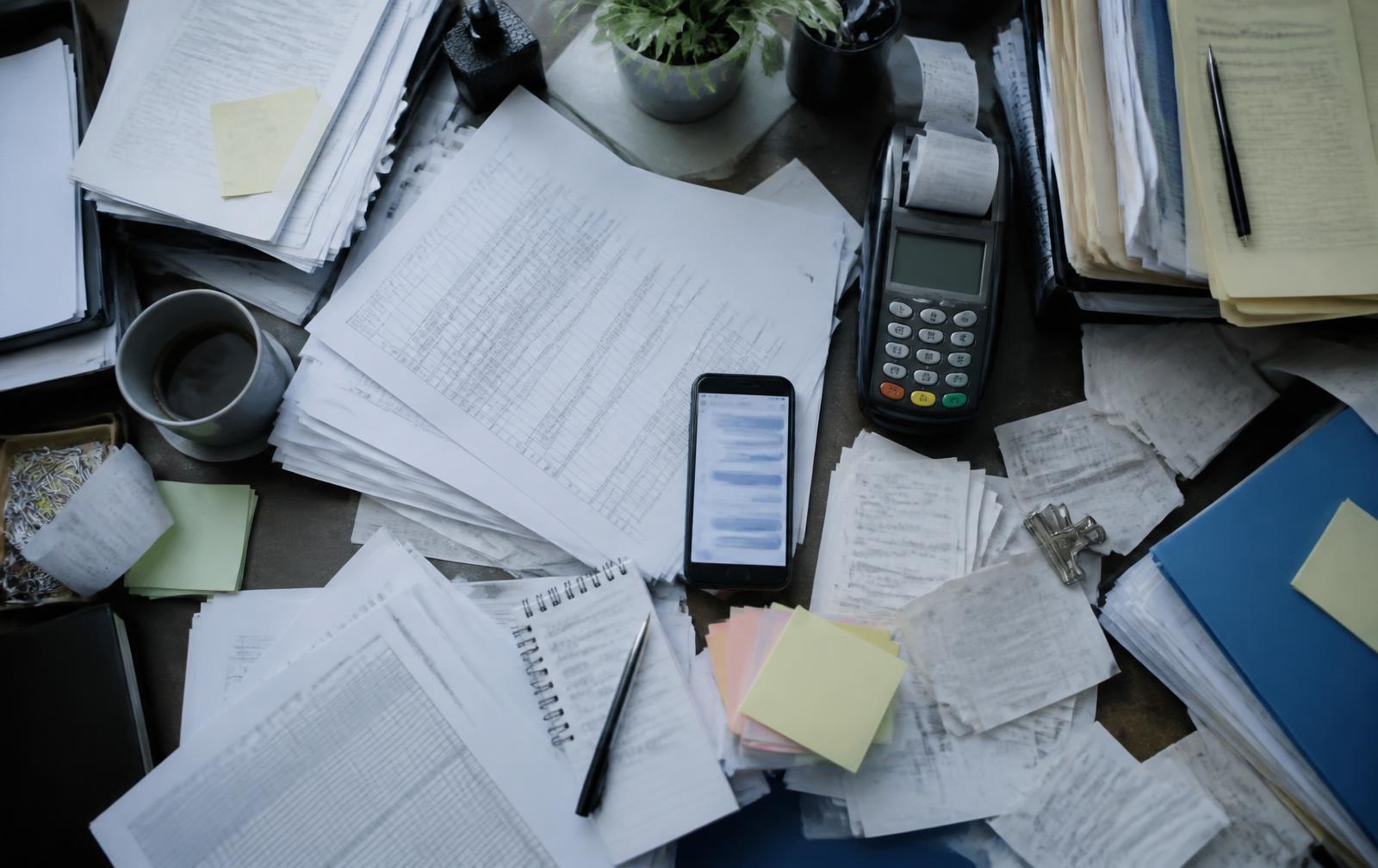 A desk covered in printed spreadsheets, receipts and a POS terminal